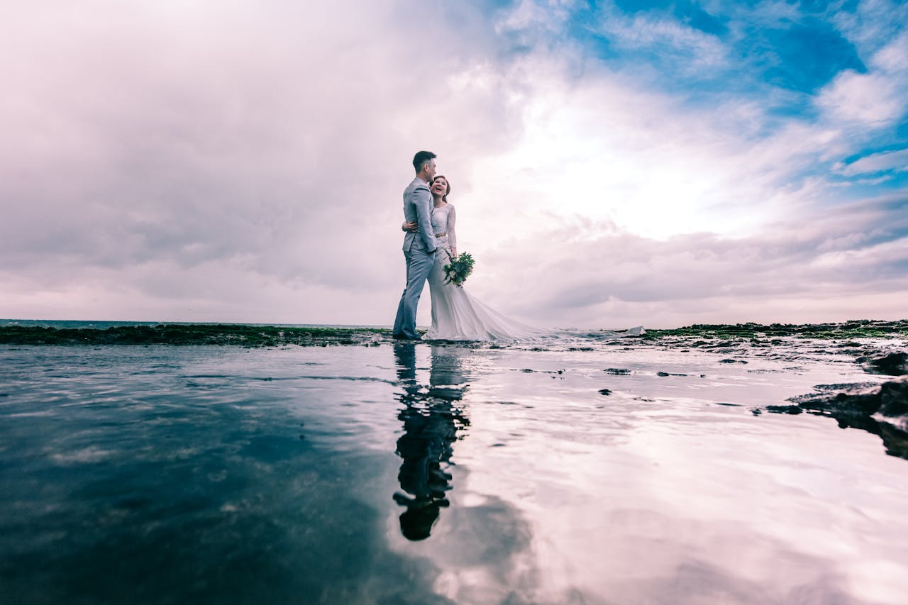 about-us-02 A joyful couple embracing on the beach in wedding attire under a dramatic sky.